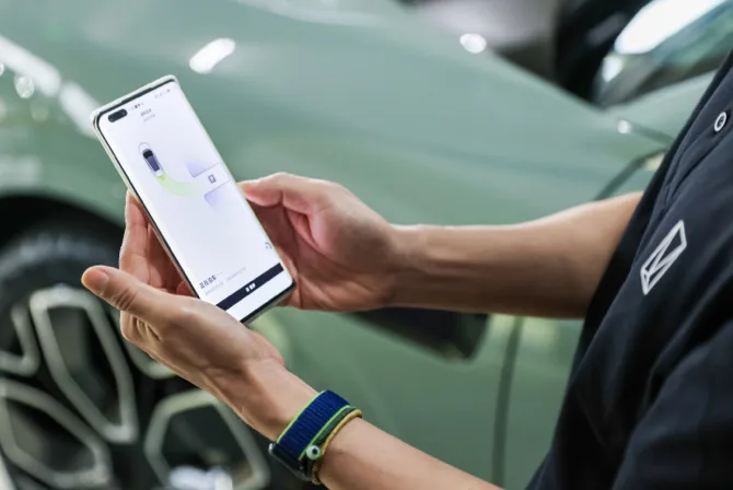 Detal - ręka trzymająca smartfon z aplikacją do zdalnej obsługi Avatr 11, w tle rozmyta zielona karoseria pojazdu