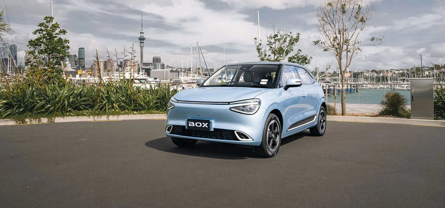 Dongfeng Box w jasnoniebieskim lakierze, przód i bok, na tle mariny i panoramy Auckland