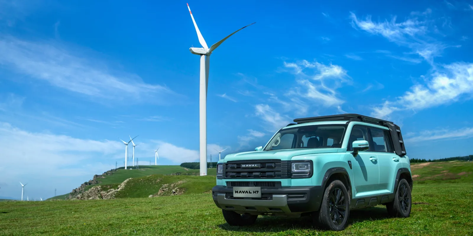 Haval H7 w kolorze miętowym, widok od przodu w 3/4, na tle farmy wiatrowej
